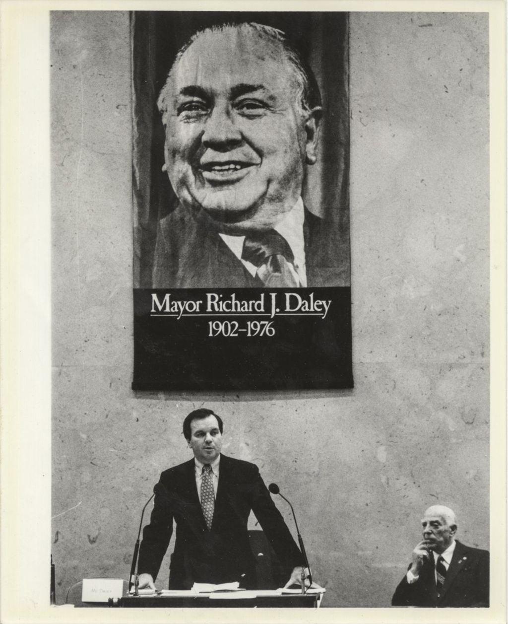 Richard M. Daley speaking in front of a memorial poster of his father
