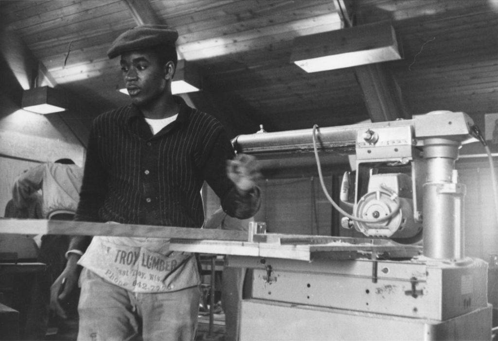 Young man standing next to a table saw in a room at the Hull-House camp in Wisconsin
