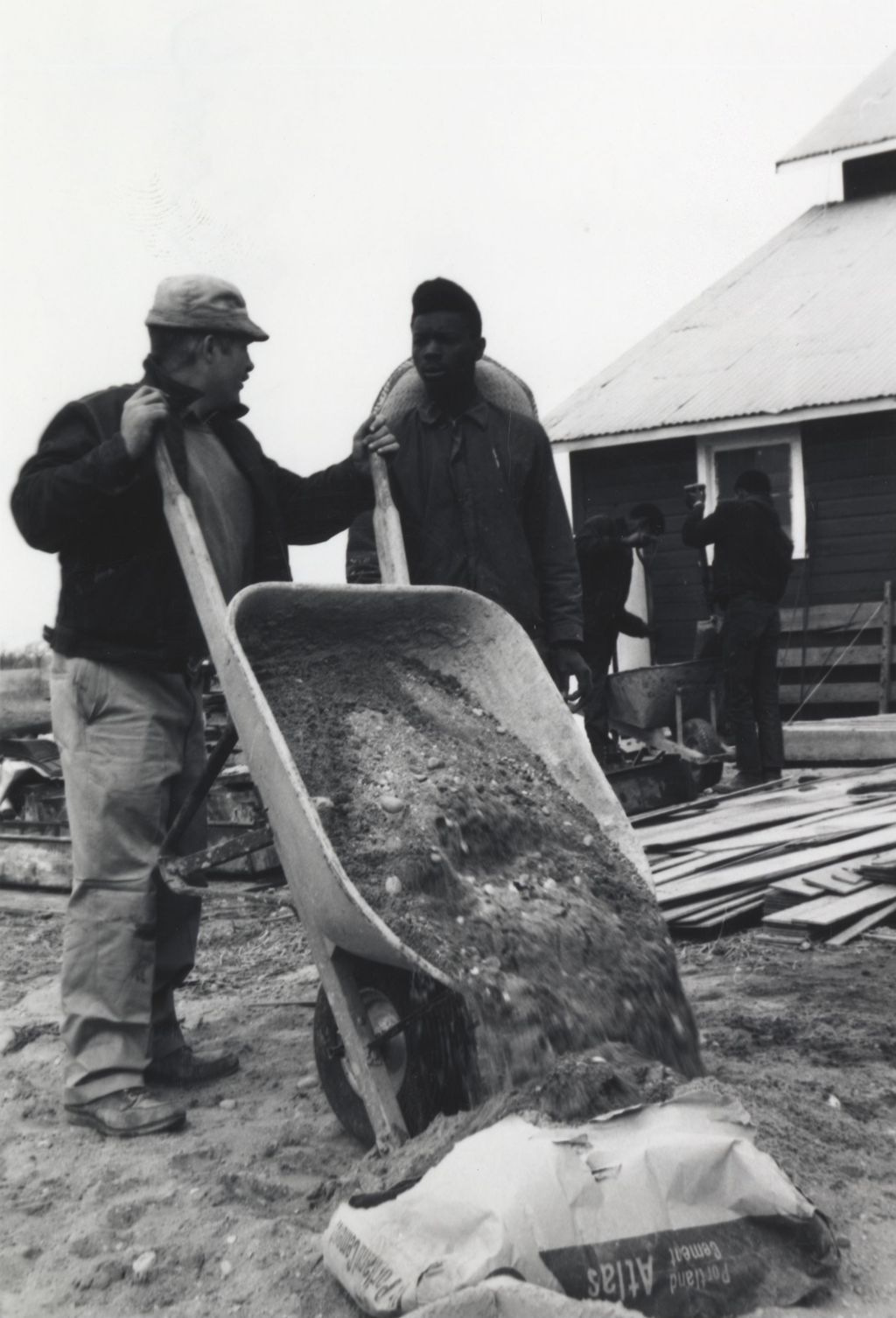 Man pours dirt out of wheelbarrow while talking to a young man at the Hull-House camp in Wisconsin