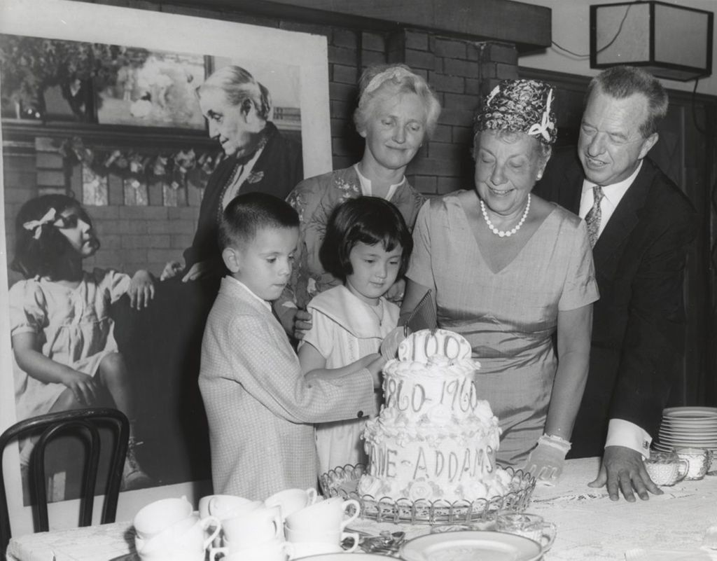 Two children cut into a cake celebrating the centennial of Jane Addams' birth; looking on are three adults: Jane Addams' grand niece, Congresswoman Marguerite Stitt Church, and the president of the Hull-House board