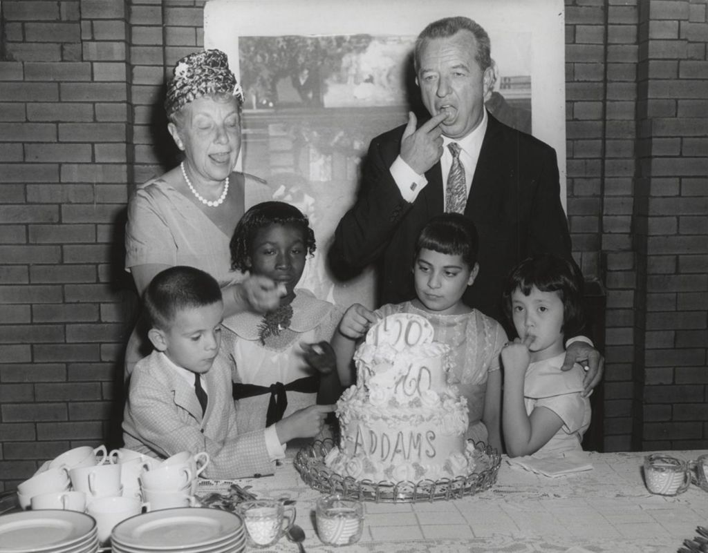 Four children, the Hull-House board president, and Congresswoman Marguerite Stitt Church use their fingers to lick frosting off a cake celebrating the centennial of Jane Addams' birth