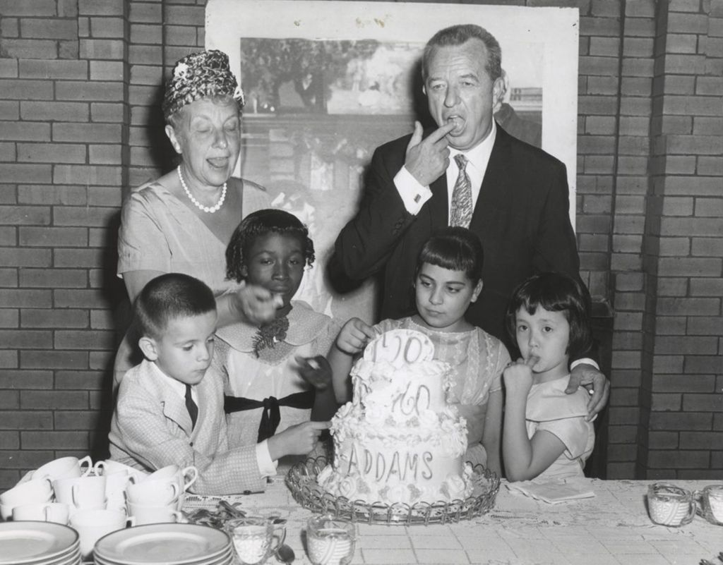 Four children, the Hull-House board president, and Congresswoman Marguerite Stitt Church use their fingers to lick frosting off a cake celebrating the centennial of Jane Addams' birth