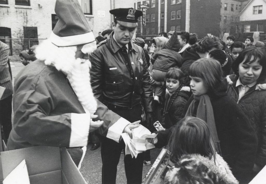 Miniature of A girl in a crowd of children hands a piece of paper to a man in a Santa Claus costume in exchange for a toy during a Hull-House Christmas giveaway