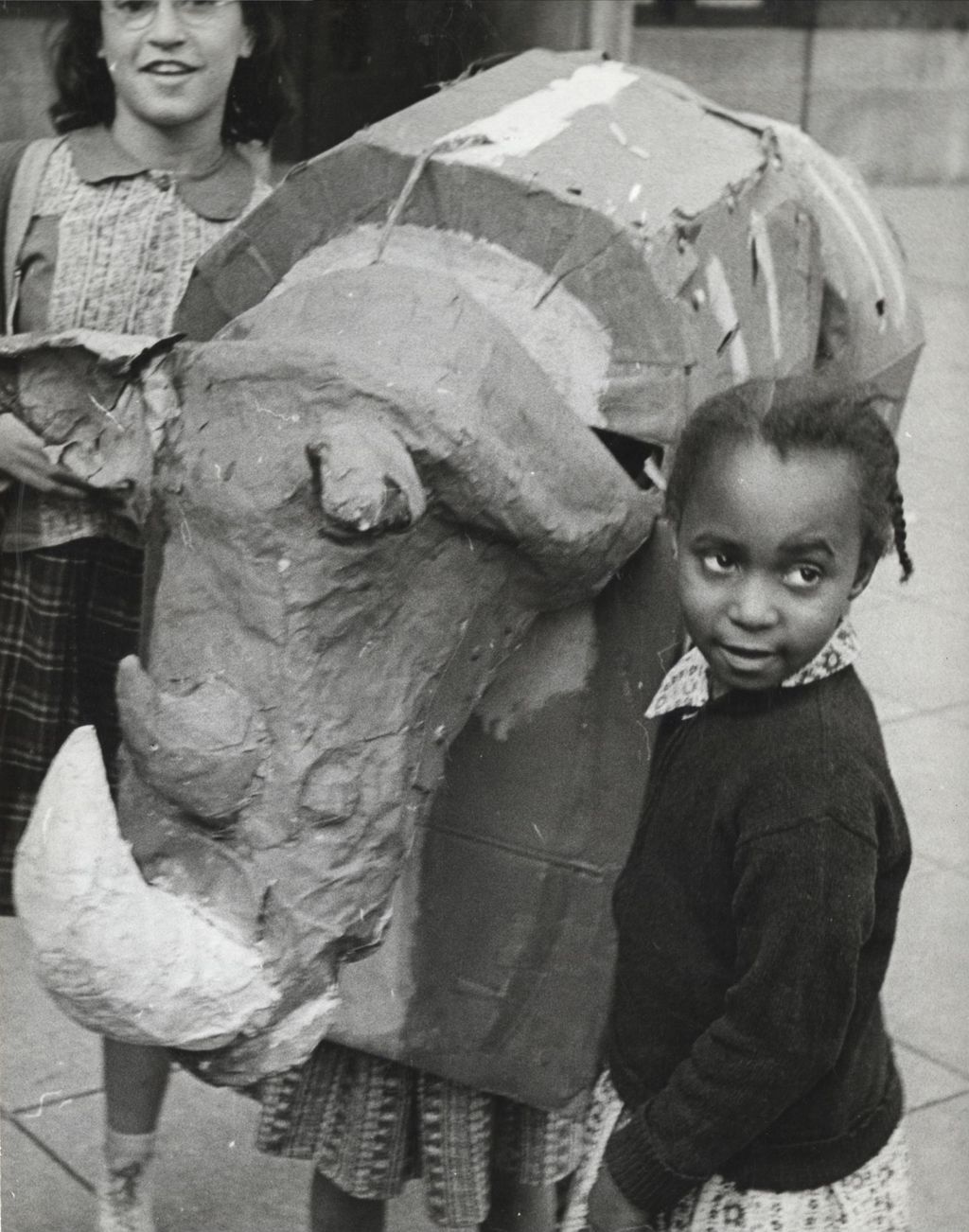 Girl standing next to girl wearing papier-mache rhinoceros costume in the Mary Crane Nursery courtyard as part of Hull-House 50th Anniversary circus