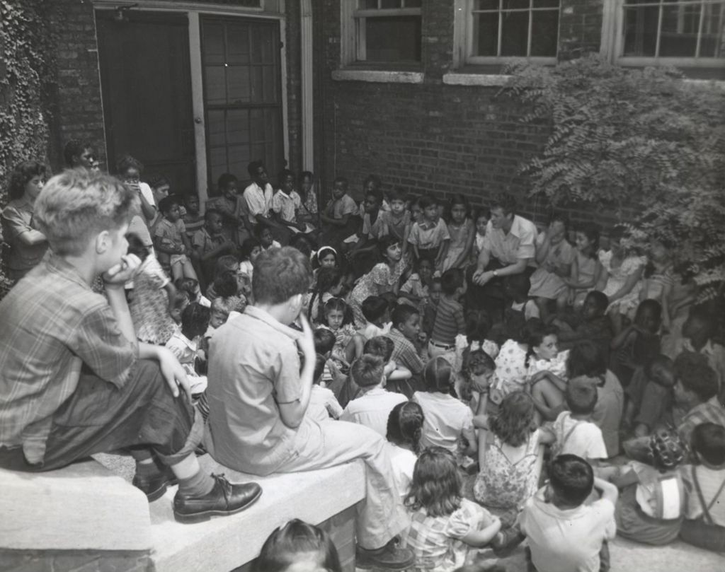 A man sits in Hull-House courtyard telling a story to dozens of children
