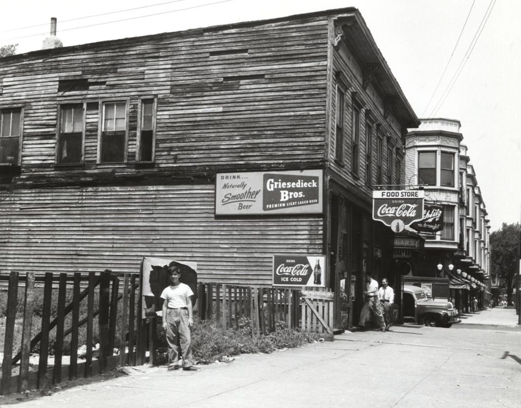 Miniature of Street scene showing young teen boy posing in front of fence next-door to deteriorating frame-built food store on Near West Side