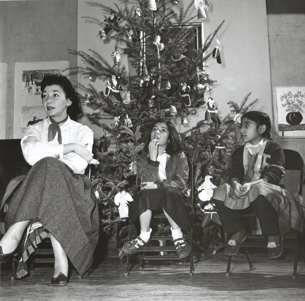 A woman and two girls sit on folding chairs in front of a Christmas tree at a Hull-House Christmas party